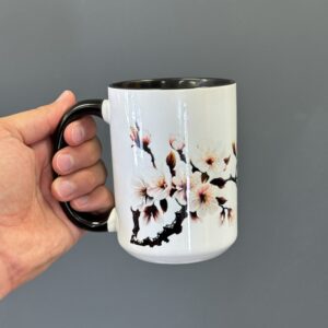 A ceramic mug featuring a cherry blossom branch design with pink and white flowers, held by a hand with a black ergonomic handle against a gray background.